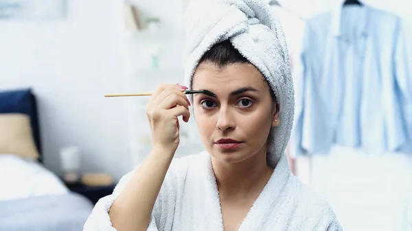 Junge Frau im Bademantel mit Kopf eingewickelt in Handtuch Styling Augenbraue mit Bürste und Blick in die Kamera im Schlafzimmer — Stockfoto