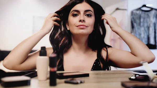 Улыбается брюнетка молодая женщина регулирует волосы с руками возле стола с декоративной косметикой в спальне — стоковое фото