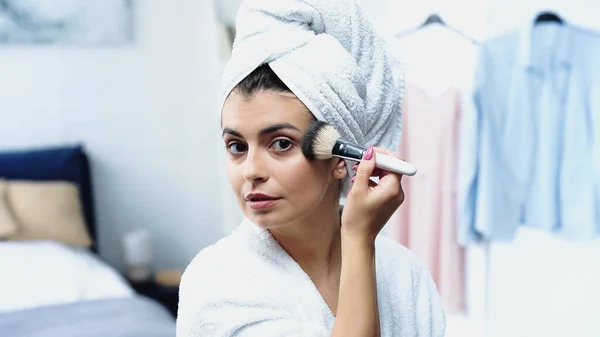 Mujer joven con la cabeza envuelta en toalla blanca aplicando polvo facial con cepillo cosmético en el dormitorio - foto de stock