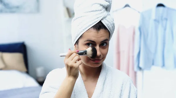 Junge Frau im Bademantel mit in Handtuch gehülltem Kopf, Gesichtspuder mit Kosmetikbürste im Schlafzimmer auftragen — Stockfoto