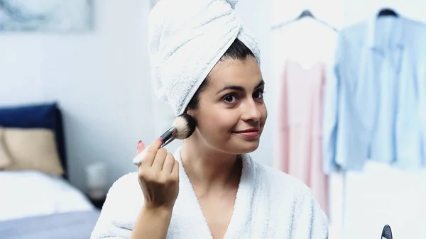 Lächelnde junge Frau mit in Handtuch gehülltem Kopf, die Gesichtspuder mit Kosmetikbürste im Schlafzimmer aufträgt — Stockfoto