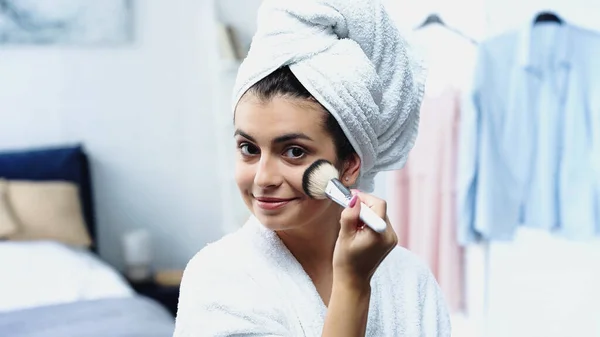 Улыбающаяся молодая женщина с головой, завернутая в белое полотенце, наносит косметический порошок на лицо в спальне — стоковое фото