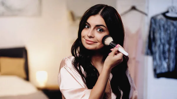 Lächelnde junge Frau in rosa Seidenpeignoir, die im Schlafzimmer Gesichtspuder mit Kosmetikbürste aufträgt — Stockfoto