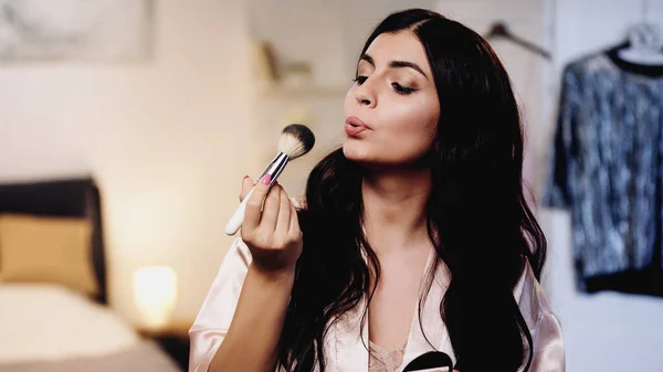 Young woman in pink silk peignoir blowing on cosmetic brush in bedroom — Stock Photo