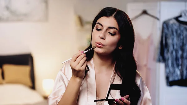 Young woman in pink silk peignoir holding eye shadows and blowing on cosmetic brush in bedroom — Stock Photo