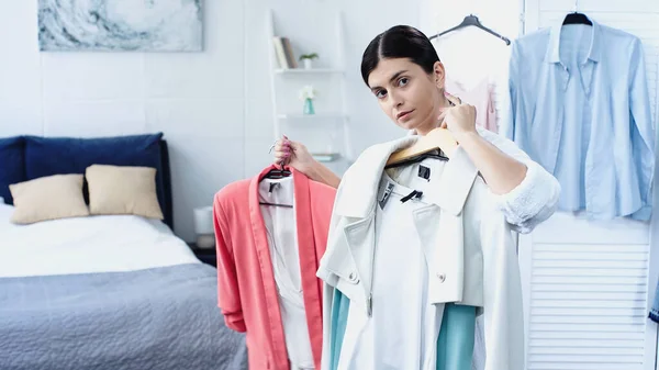 Morena mujer joven en albornoz sosteniendo la ropa en perchas en el dormitorio - foto de stock