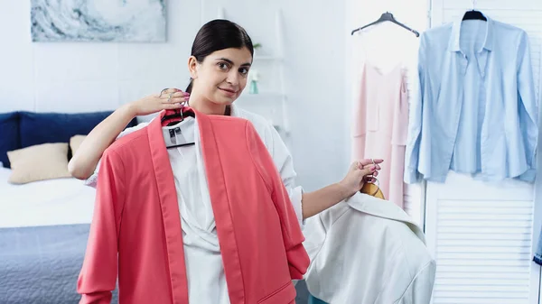 Jeune femme brune souriante en peignoir tenant des vêtements sur les cintres dans la chambre — Photo de stock