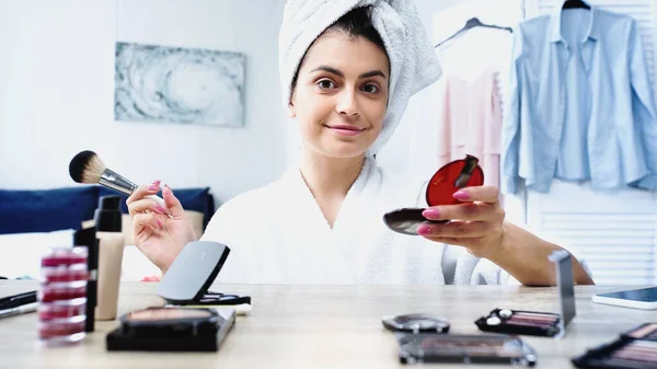 Jeune femme souriante avec la tête enveloppée dans une serviette tenant brosse cosmétique et poudre visage dans la chambre — Photo de stock