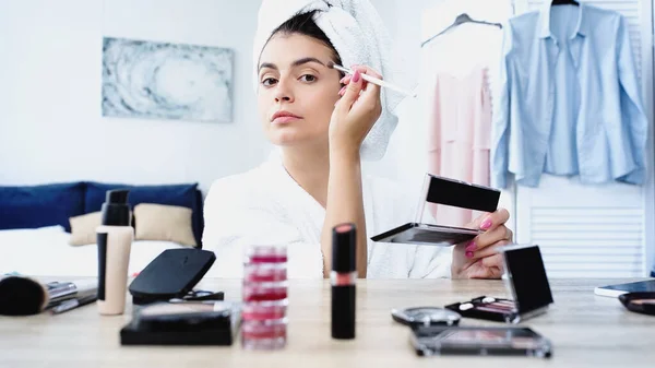 Mujer joven en albornoz aplicando sombras de ojos con cepillo cosmético cerca de la mesa con cosméticos decorativos en el dormitorio - foto de stock