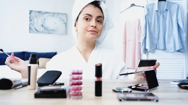 Fröhliche junge Frau im Bademantel sitzt mit Kosmetikbürste und Lidschatten am Tisch mit dekorativer Kosmetik im Schlafzimmer — Stockfoto