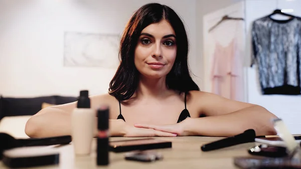 Jeune femme brune souriante aux cheveux brillants assise près de la table avec des cosmétiques décoratifs dans la chambre — Photo de stock