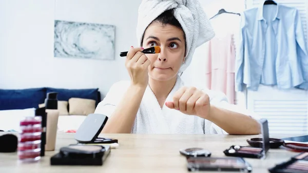 Jovem com cabeça embrulhada em toalha grimacing e aplicando fundação rosto com escova cosmética no quarto — Fotografia de Stock
