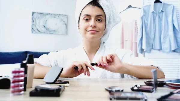 Giovane donna sorridente in accappatoio bianco con testa avvolta in asciugamano seduta vicino al tavolo con cosmetici decorativi in camera da letto — Foto stock