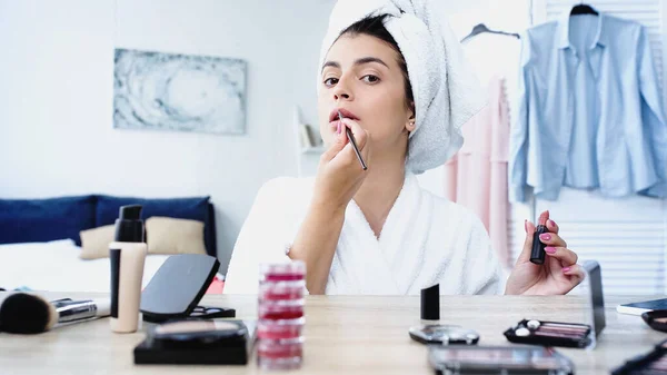 Young woman applying lipstick with cosmetic brush near table with decorative cosmetics in bedroom — Stock Photo