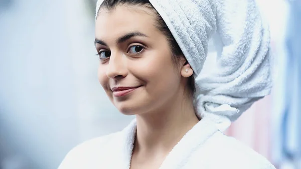 Jeune femme souriante en peignoir avec la tête enveloppée dans une serviette en regardant la caméra dans la chambre — Photo de stock