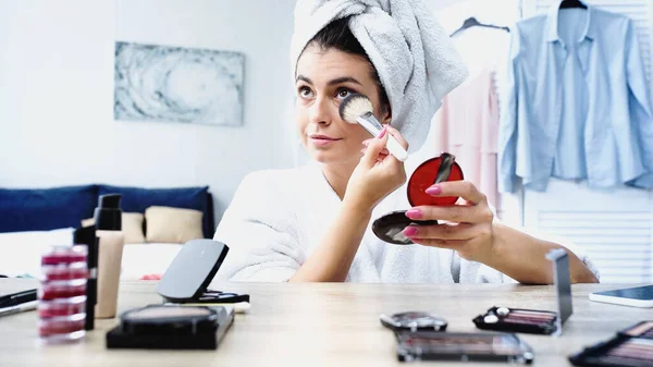 Jeune femme en peignoir avec tête enveloppée dans une serviette appliquer la poudre visage avec pinceau cosmétique dans la chambre — Photo de stock