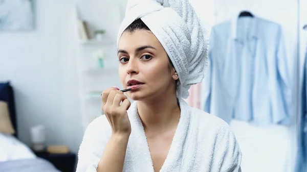 Mujer joven en albornoz con cabeza envuelta en toalla aplicando lápiz labial con cepillo cosmético en el dormitorio - foto de stock