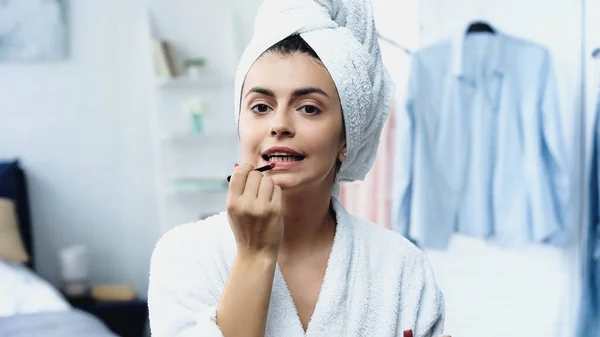 Jovem mulher em roupão de banho com cabeça embrulhada em toalha aplicando batom com escova cosmética e olhando para a câmera no quarto — Fotografia de Stock