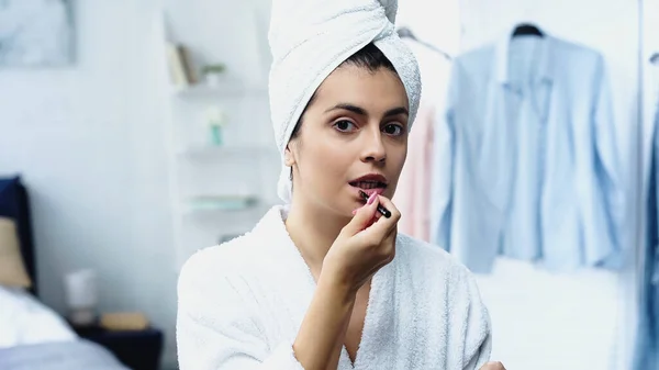 Junge Frau im Bademantel mit Kopf in Handtuch gehüllt Lippenstift mit Kosmetikbürste im Schlafzimmer auftragen — Stockfoto