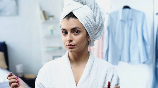 Jeune femme en peignoir avec la tête enveloppée dans une serviette tenant rouge à lèvres et pinceau cosmétique dans la chambre — Photo de stock