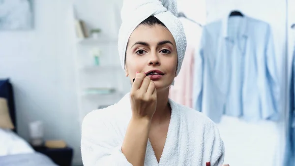 Jovem mulher em roupão de banho com cabeça embrulhada em toalha aplicando batom com escova cosmética no quarto — Fotografia de Stock