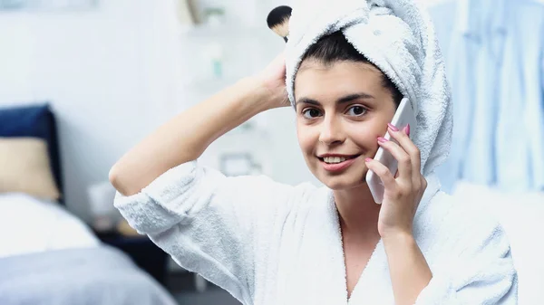 Young woman with head wrapped in towel holding cosmetic brush and speaking on cellphone in bedroom — Stock Photo