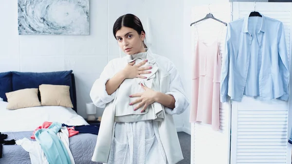 Young woman in bathrobe holding jacket near bed with clothes in bedroom — Stock Photo