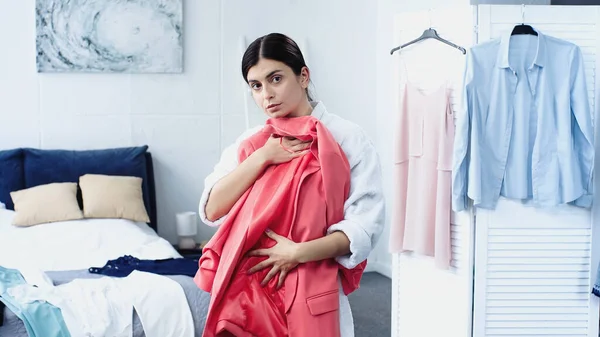 Young woman in bathrobe holding pink blazer near bed with clothes in bedroom — Stock Photo