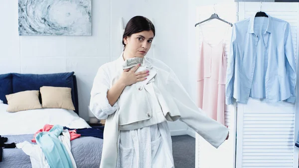 Young woman in white bathrobe fitting jacket near bed with clothes in bedroom — Stock Photo