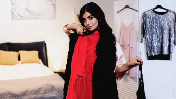 Young woman in pink silk peignoir trying on dresses on hangers in bedroom — Stock Photo