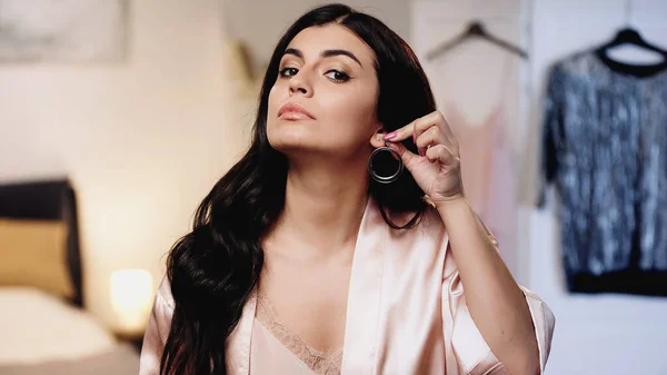 Young woman in pink silk peignoir trying on earrings in bedroom — Stock Photo