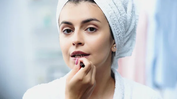 Portrait of young woman with head wrapped in towel applying lipstick with cosmetic brush in bedroom — Stock Photo