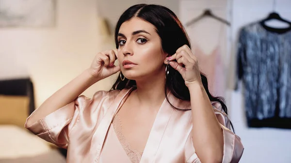 Young woman in silk peignoir trying on earrings in bedroom — Stock Photo