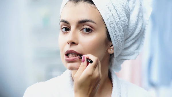 Porträt einer jungen Frau mit in Handtuch gehülltem Kopf, die Lippenstift mit Kosmetikbürste im Schlafzimmer aufträgt — Stockfoto