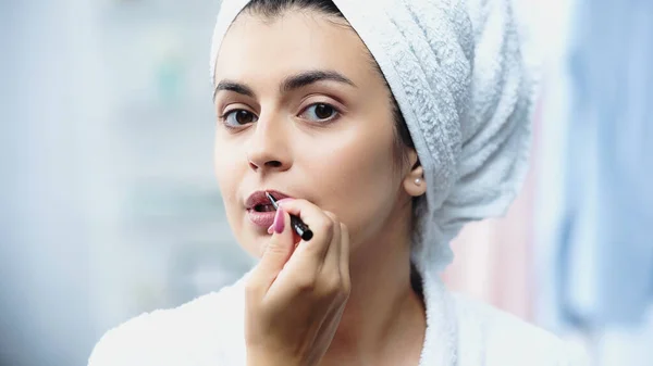 Porträt einer jungen Frau mit in Handtuch gehülltem Kopf, die Lippenstift mit Kosmetikbürste im Schlafzimmer aufträgt — Stockfoto