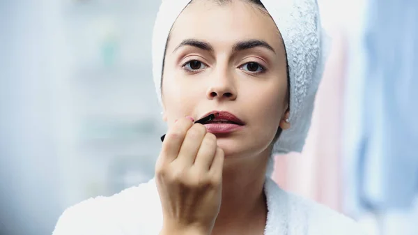 Portrait of young woman with head wrapped in white towel applying lipstick with cosmetic brush in bedroom — Stock Photo