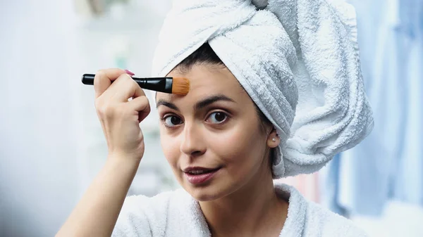 Portrait of young woman with head wrapped in towel applying foundation on forehead in bedroom — Stock Photo