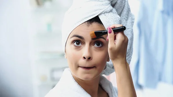 Porträt einer jungen Frau mit in Handtuch gehülltem Kopf, die mit Kosmetikbürste im Schlafzimmer Grimassen schlägt und Foundation auf das Gesicht aufträgt — Stockfoto