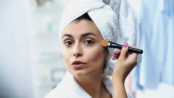 Jovem com cabeça embrulhada em toalha aplicando fundação no rosto com escova cosmética no quarto — Fotografia de Stock