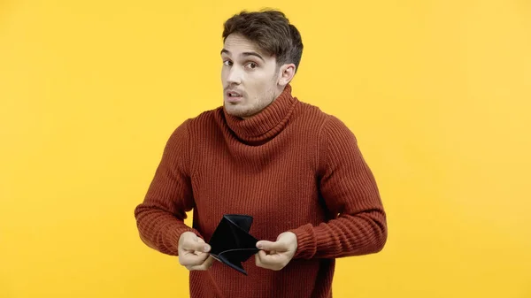 Запутавшийся мужчина держит пустой бумажник и смотрит на камеру, изолированную на желтый — стоковое фото