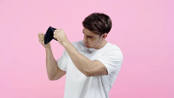 Hombre joven en camiseta blanca mirando a la cartera vacía aislado en rosa — Stock Photo
