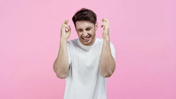 Homem sorridente com olhos fechados mostrando dedos cruzados isolados em rosa — Fotografia de Stock