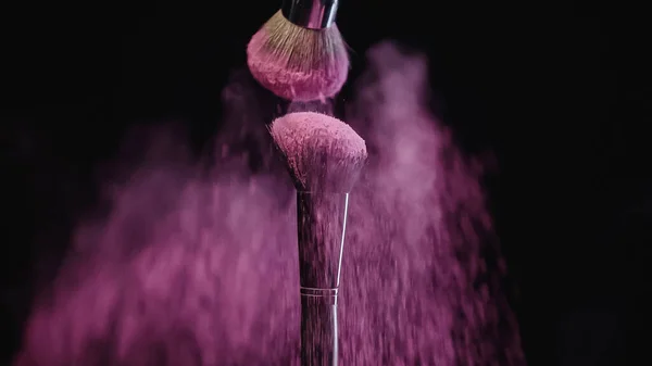 Cepillos cosméticos con polvo de holi rosa cayendo sobre negro - foto de stock