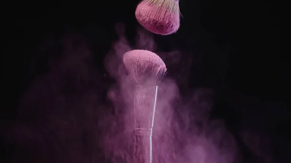 Spazzole cosmetiche morbide vicino spruzzi di polvere rosa su sfondo nero — Foto stock