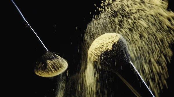 Cosmetic brushes with yellow powder hitting and making splash of dust on black background — Stock Photo