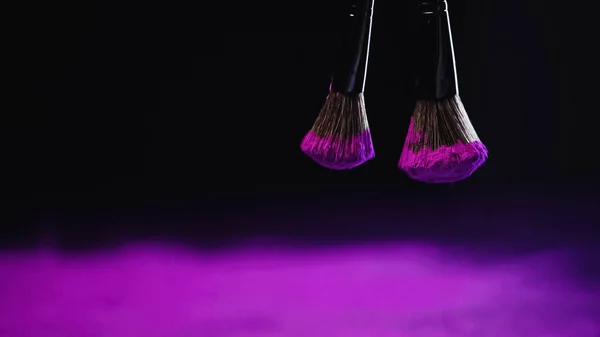 Cosmetic brushes with purple holi powder hanging on black background — Stock Photo