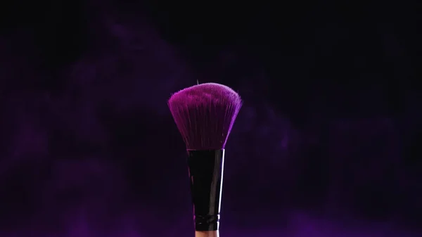 Cosmetic brush with purple holi powder near dust on black background — Stock Photo