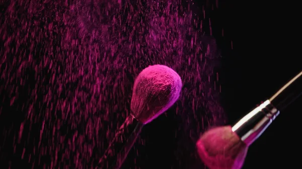 Cosmetic brushes with pink holi powder near dust splashing on black background — Stock Photo