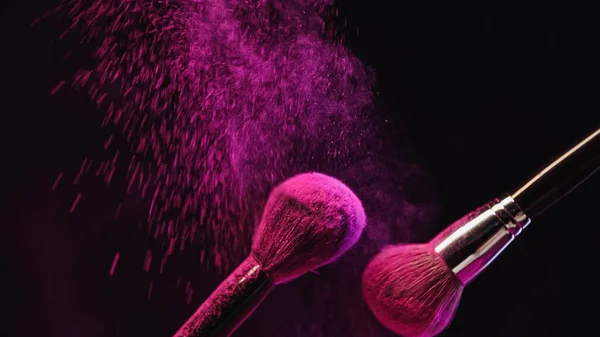 Cosmetic brushes with bright pink powder splashing on black background — Stock Photo