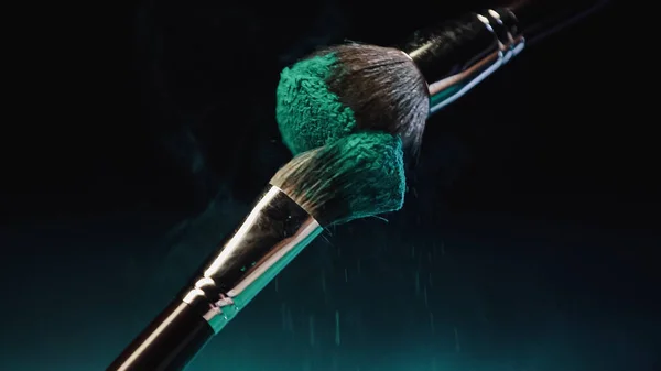 Cosmetic brushes with vibrant turquoise holi paint near dust on black background — Stock Photo